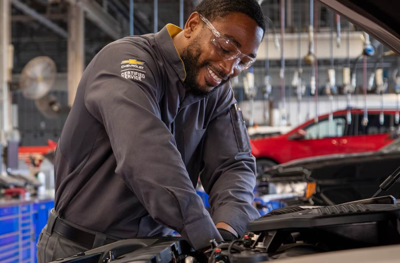 Chevrolet mechanic wearing safety glasses working under an open car hood in a service bay.